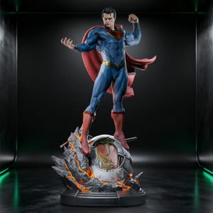 Könnte beinhalten: Eine detaillierte Superman-Statue, die den Charakter in einem blauen Anzug mit rotem Umhang und gelbem Gürtel zeigt, stehend auf einem Sockel aus gezacktem, dunkelgrauem Fels mit leuchtend orangefarbenen Lava-Akzenten und einer eingebetteten Metallkugel.