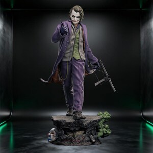 May include: A detailed collectible figure of a character with white face paint, red lipstick, and messy hair. The figure wears a long purple coat, a green vest, and purple trousers. It holds a small knife and a submachine gun, standing on a rocky base.