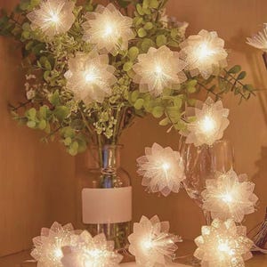 Puede incluir: Una guirnalda de luces LED con forma de flores blancas de múltiples pétalos y texturas brillantes. Las luces están dispuestas entre follaje verde en un florero de vidrio transparente, emitiendo una luz cálida desde el centro de cada flor.