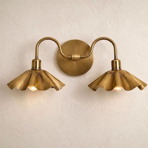 May include: A two-light wall sconce featuring a circular brass backplate and two curved arms extending to support downward-facing, ruffled, bell-shaped brass shades. Each shade houses a single light bulb that emits a warm, soft glow.