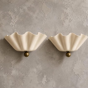 Op de afbeelding: Een paar gebroken witte wandlampen met een breed, golvend, schelpachtig kapontwerp. Elke lamp is aan de basis bevestigd met een klein, bolvormig metalen accentstuk in antieke messingkleur, wat contrasteert met de gladde, gebogen keramische vorm.