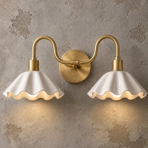 Puede incluir: Un aplique de pared doble con una placa circular de latón y dos brazos curvos de latón que sostienen dos pantallas de cerámica blanca, acanaladas y en forma de campana. La lámpara emite un brillo cálido bajo cada pantalla, resaltando el metal.