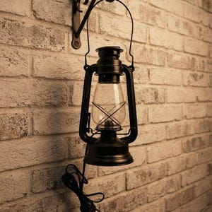 May include: A black metal lantern wall lamp featuring a clear glass chimney and a wire cage guard. The lantern hangs from a decorative black metal wall bracket and includes a black electrical cord with a plug extending from the base of the lamp.