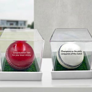 Pode incluir: Duas bolas de críquete, uma vermelha e uma branca, cada uma numa caixa expositora quadrada transparente com base de relva artificial verde. A bola vermelha tem o texto Congratulation Tom For your Great Victory, e a branca Champions on the pitch, conquerors of the match.