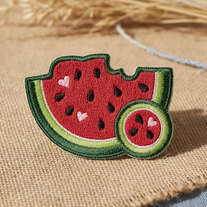 May include: An embroidered patch shaped like a watermelon slice with a bite taken out of it and a small circular watermelon piece. The design features red fruit, black seeds, light pink heart-shaped seeds, and a green rind with a light green border.