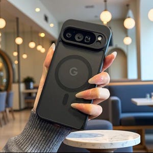 May include: A matte black smartphone case featuring a raised camera bar with multiple lenses and a circular magnetic charging ring on the back. The centre of the case displays a small, circular Google G logo, and the design includes a vertical alignment magnet below the ring.
