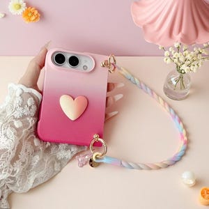 May include: A phone case featuring a pink and light pink gradient finish with a central heart-shaped emblem. The case includes a detachable, braided, multi-colored pastel strap attached by gold-toned metal rings and a small bear-shaped charm.