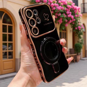 Puede incluir: Una funda de smartphone negra con acabado brillante, bordes metálicos dorados y protector de lente de cámara. La parte trasera muestra un icono de corazón, el texto "Smile every day" y un soporte de anillo magnético circular. El lateral está decorado con pequeños corazones y la palabra "nice."
