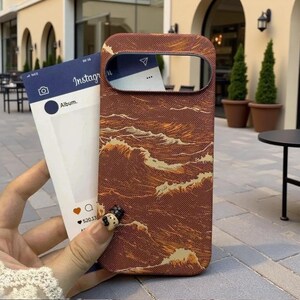 May include: A brown smartphone case featuring a textured pattern of stylized golden waves. The case has a large horizontal cutout for the camera module. Behind the case is a white card printed with an Instagram interface, including the text Album.