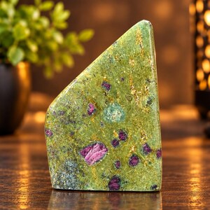 May include: A polished, wedge-shaped piece of zoisite mineral featuring a vibrant green base colour with embedded deep magenta ruby crystals and subtle golden-brown mineral inclusions throughout the smooth, flat, and angular stone surface.