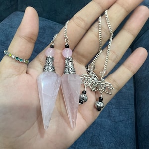 May include: Two rose quartz pendulums with silver-toned metal caps featuring intricate scrollwork. Each pendulum is topped with a small pink bead and a black bead. A silver-toned chain with a lotus flower charm and clear beads is attached to the top.