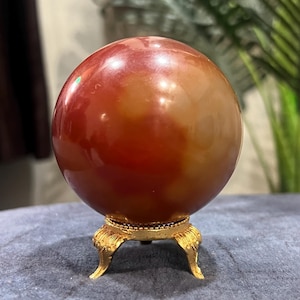 Peut inclure: Une pierre sphérique lisse et polie présentant un dégradé de tons rouge-orangé profond et ambré translucide. La sphère repose solidement sur un petit support en métal doré orné, doté de trois pieds incurvés aux détails décoratifs.