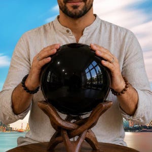 May include: A large, smooth, polished black sphere made of obsidian stone, resting on a carved wooden stand with an intricate, interlocking base design. The surface of the sphere is highly reflective, showing a clear mirror-like image of surroundings.