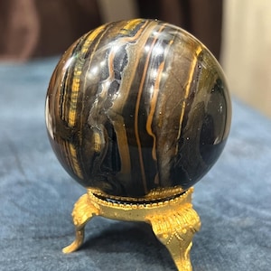 May include: A polished tiger's eye stone sphere featuring deep black and golden-brown banded patterns. The smooth, reflective orb is supported by a decorative gold-toned metal stand with three ornate, curved legs and a textured circular base.