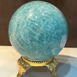 May include: A polished, spherical amazonite gemstone featuring a vibrant teal-green colour with intricate white veining throughout. The stone is securely displayed on a small, ornate gold-toned metal stand with three curved legs and a beaded rim.