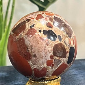 Peut inclure: Une pierre sphérique polie présentant un motif conglomérat de fragments rouges, bruns, noirs et crème incrustés dans une matrice. La sphère repose sur un petit support en métal doré orné, doté de trois pieds incurvés et de bases décoratives.