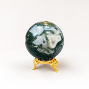 May include: A polished moss agate sphere featuring deep forest green tones and translucent white inclusions, resting on a small, ornate gold-coloured metal tripod stand with curved legs and detailed decorative feet.