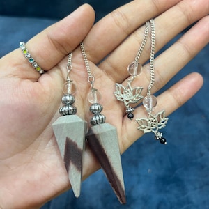 May include: Two pointed stone pendulums with grey and brown patterns, each featuring a silver-toned metal bead, a clear spherical bead, and a silver-toned chain. Two additional silver-toned lotus flower charms are attached to the chains.