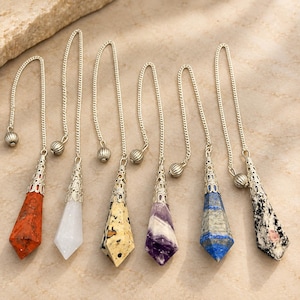 Puede incluir: Seis péndulos de piedras preciosas en forma de punta que cuelgan de cadenas de metal plateado. Cada péndulo presenta una piedra única, incluyendo jaspe rojo, cuarzo blanco, jaspe moteado, amatista, lapislázuli azul y granito blanco y negro.