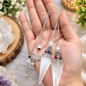 May include: Two clear, pointed crystal pendulums suspended from silver-toned chains. Each pendulum features a decorative metal cap with small red and blue accents, topped with a white bead. A silver lotus charm hangs from one chain.