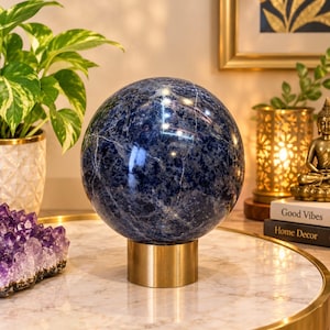 Può includere: Una sfera lucida di colore blu intenso con venature bianche, poggiata su un supporto cilindrico in metallo dorato. La sfera presenta una superficie liscia e riflettente con inclusioni minerali naturali e una tonalità blu navy e reale.