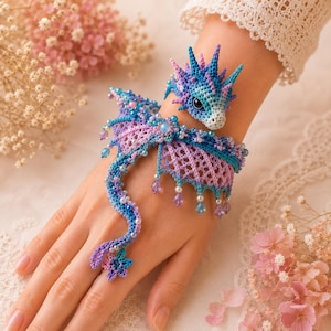 May include: A crochet dragon bracelet featuring a blue and purple color palette with intricate wing details, small beads, and a dragon head design that wraps around the wrist, extending down to a small star-shaped ring on the finger.