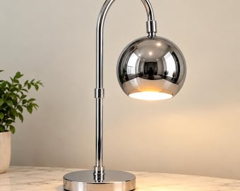 Puede incluir: Una lámpara de mesa de cromo pulido con un cuello delgado y curvado y una pantalla esférica reflectante. La lámpara tiene una base circular y emite una luz cálida dirigida hacia abajo desde la abertura inferior del cabezal metálico.