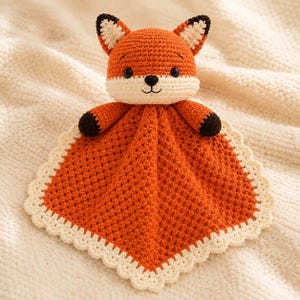 Puede incluir: Una manta de seguridad de zorro tejida a ganchillo con una cabeza de zorro rellena de hilo naranja y blanco, ojos de seguridad negros y nariz marrón oscuro. El cuerpo de la manta es de hilo naranja texturizado con un borde festoneado crema.