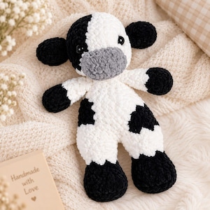 May include: A plush cow toy crafted from soft, textured yarn in white and black. The stuffed animal features a gray snout, round black ears, and black patches on its body and limbs. It has two small, dark, glossy eyes set into the white head.
