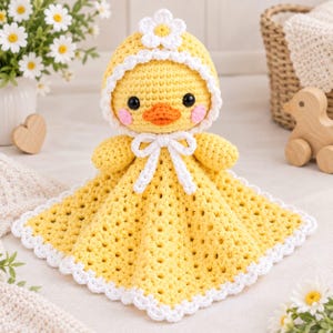 Pode incluir: Uma manta de segurança de crochê amarela com uma cabeça de pato, um capuz branco, uma flor branca no topo e um laço branco. O pato tem olhos pretos, um bico laranja e bochechas rosadas. A manta possui uma borda branca com acabamento festonado.
