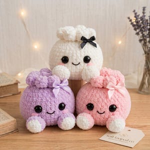 Può includere: Tre peluche rotondi all'uncinetto con grandi occhi neri, guance rosa e piccoli sorrisi. Il giocattolo in alto è bianco con un fiocco nero, quello in basso a sinistra è color lavanda con un fiocco viola e quello in basso a destra è rosa chiaro con un fiocco rosa chiaro.
