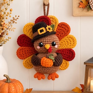 Puede incluir: Un adorno de pared de pavo tejido a ganchillo con cuerpo marrón, un abanico de plumas amarillas, naranjas y rojas, y un sombrero negro con hebilla amarilla y girasol. El pavo sostiene una pequeña calabaza naranja y tiene patas de color naranja.