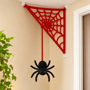 Op de afbeelding: Een rode gehaakte spinnenweb hoekdecoratie bevestigd aan de muur met witte zelfklevende haken. Een enkel rood gehaakt koord hangt vanuit het midden van het web en houdt een zwarte gehaakte spin vast met grote, ronde, donkere ogen.