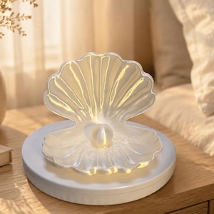 May include: A translucent, white, shell-shaped lamp featuring a central, spherical light element that mimics a pearl. The shell is open, revealing the glowing orb inside, and it is mounted on a circular, white base that provides a stable foundation.