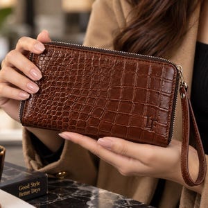 May include: A rectangular brown wallet with a textured crocodile skin pattern, a silver-toned metal zip closure, and a matching wrist strap. The bottom right corner features the embossed letters LF. The wallet has a structured, slim design.