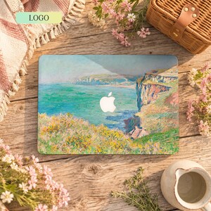 Può includere: Una skin rettangolare per computer portatile con un dipinto impressionista di un paesaggio costiero con acqua turchese, scogliere scoscese e fiori di campo gialli. Il logo Apple bianco è centrato sulla superficie della skin del portatile.