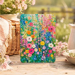 Puede incluir: Una funda rectangular para tableta con un estampado floral multicolor y vibrante. El diseño muestra un denso prado de flores silvestres en tonos rosa, naranja, amarillo, azul y blanco sobre un fondo verde, cubriendo toda la superficie.