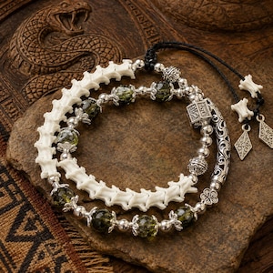 Könnte beinhalten: Ein mehrsträngiges Armband mit einer Reihe von weißen, knochenartigen Wirbelsegmenten, durchscheinenden olivgrünen Perlen und silberfarbenen Metallabstandshaltern. Das Design umfasst einen quadratischen gravierten Anhänger und zwei rautenförmige Metallanhänger.