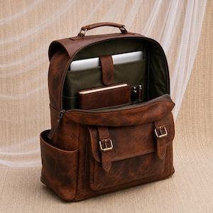 May include: A brown leather backpack with a top handle and a front pocket secured by two brass-toned buckles. The interior features a dark green lining, a dedicated laptop sleeve with a strap, a notebook, and two pen holders inside the main compartment.