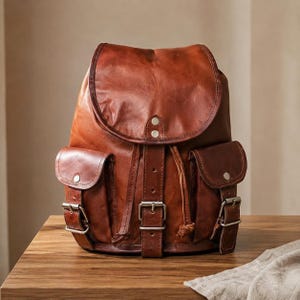 Può includere: Uno zaino in pelle marrone medio caratterizzato da una grande patta superiore con due chiusure a scatto argentate, due tasche laterali con cinturini con fibbia e un cinturino verticale centrale con fibbia in metallo. Include una chiusura a coulisse.