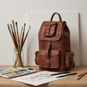 May include: A brown leather backpack featuring a top handle, a flap with a buckle closure, a front pocket with a buckle strap, and two side pockets. The backpack is positioned next to a glass jar filled with various paintbrushes and a paint palette.
