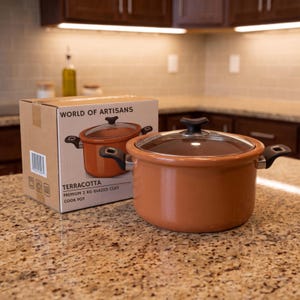 May include: A terracotta-colored glazed clay cook pot with a clear glass lid and black handles, positioned next to its cardboard packaging labeled World of Artisans, Terracotta Premium 2 kg Glazed Clay Cook Pot, featuring a barcode and icons.