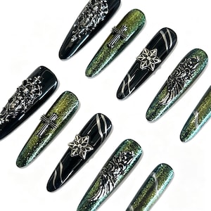 May include: A set of ten press-on nails featuring a mix of glossy black and shimmering green cat-eye finishes. The nails are adorned with silver-toned metal charms including crosses, stars, and ornate filigree wing designs in various shapes.