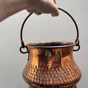 Könnte beinhalten: Ein runder Kupfertopf mit gehämmertem Muster auf der unteren Hälfte, ausgestattet mit drei kurzen Beinen und einem dünnen, dunklen Metallhenkel, der an zwei seitlichen Ringen befestigt ist. Die Oberfläche zeigt ein poliertes Finish mit Patina.