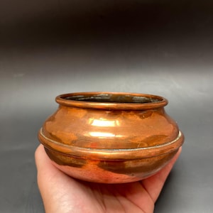 Könnte beinhalten: Eine kleine, runde Schale aus poliertem Kupfer mit einer glatten, reflektierenden Oberfläche und einem leicht ausgestellten Rand. Die Schale weist eine ausgeprägte horizontale Rille in der Mitte auf, die eine zweigeteilte Optik erzeugt.
