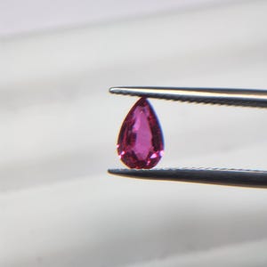 以下が含まれることがあります： 鮮やかな濃いピンク色のペアシェイプ（洋梨型）のファセットカットされた宝石。ピンセットのギザギザした金属製の先端でしっかりと挟まれており、その半透明の質感と精密なカットがはっきりと見て取れます。