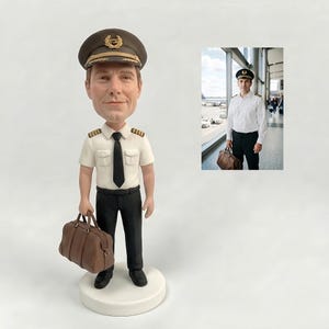May include: A custom bobblehead figurine depicting a pilot wearing a white short-sleeved shirt with epaulets, a black tie, black trousers, and a dark brown captain's hat, holding a brown leather-style travel bag, standing on a circular white base.
