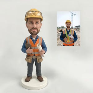 May include: A custom bobblehead figurine of a construction worker with a beard, wearing a yellow hard hat, safety glasses, an orange high-visibility vest, a blue shirt, and gray pants. The figure holds a tablet and a rolled-up white blueprint.