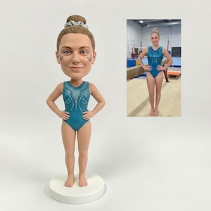 May include: A custom bobblehead figurine of a gymnast with brown hair in a bun, wearing a teal leotard with silver embellishments and small butterfly decals on the cheeks. The figure stands on a round white base, mirroring a photograph of a gymnast.