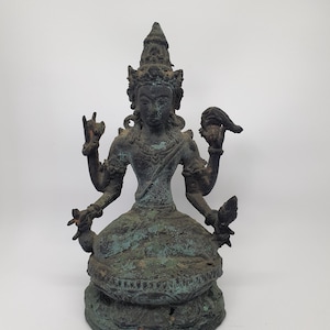 May include: A dark bronze-coloured metal statue of a four-armed deity seated in a cross-legged position on a lotus base. The figure wears an ornate headdress and jewellery, with a weathered, oxidised patina showing patches of green and brown throughout.